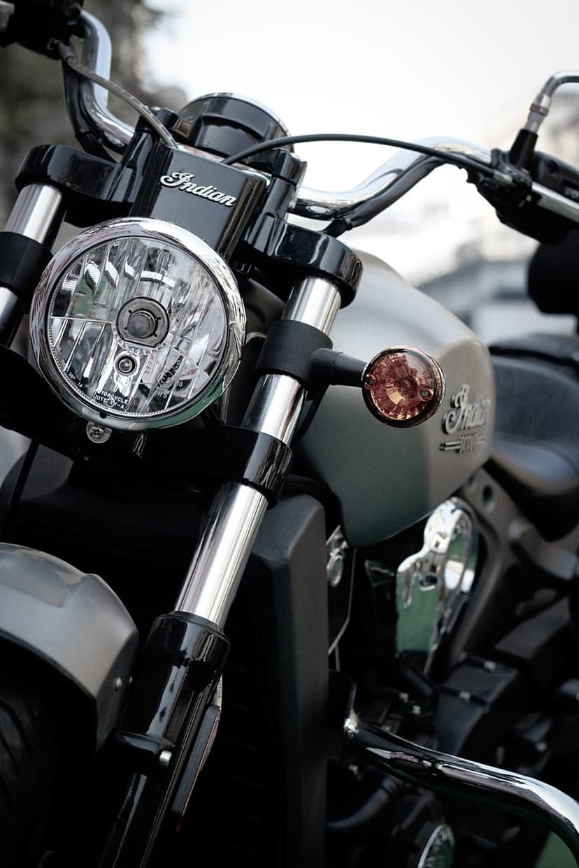 Chrome handlebars and a round headlight of an Indian motorcycle with a silver logo on the black fuel tank.