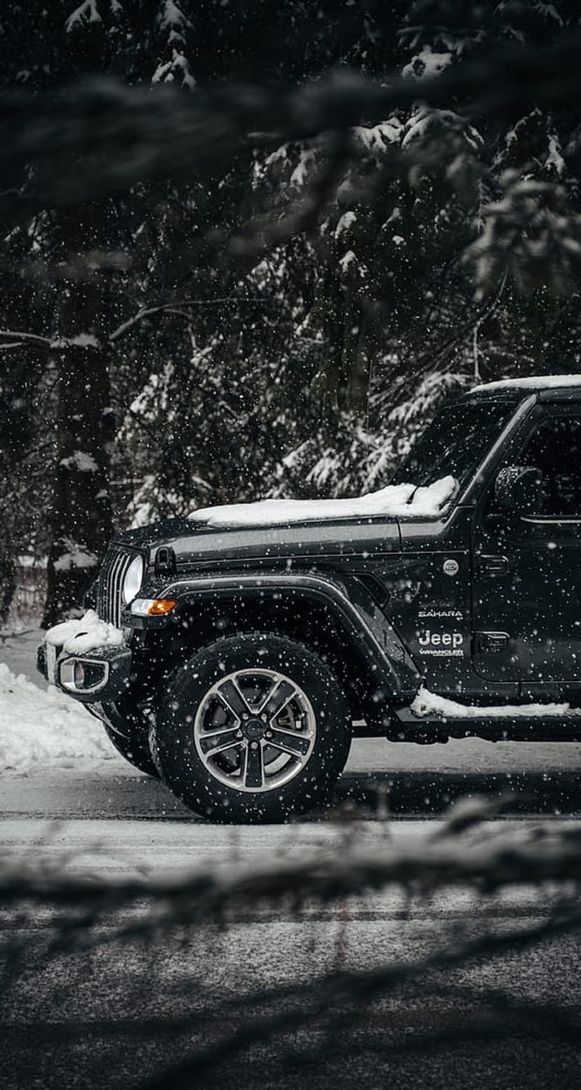 Black Jeep Wrangler on a snow-covered forest road with falling snowflakes and frozen pine trees in the background.