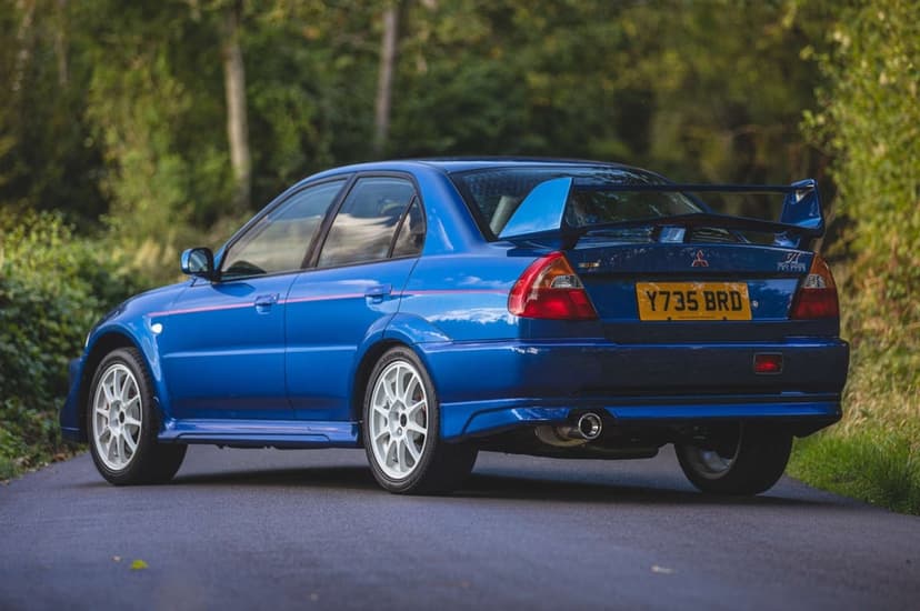 Rear view of a blue Mitsubishi Lancer Evolution VI with white wheels and a large wing on a paved forest road.