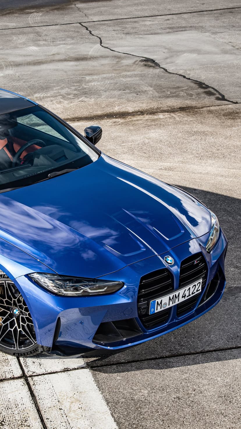 Metallic blue BMW M4 Competition on gray asphalt with large kidney grille and carbon fiber mirrors.
