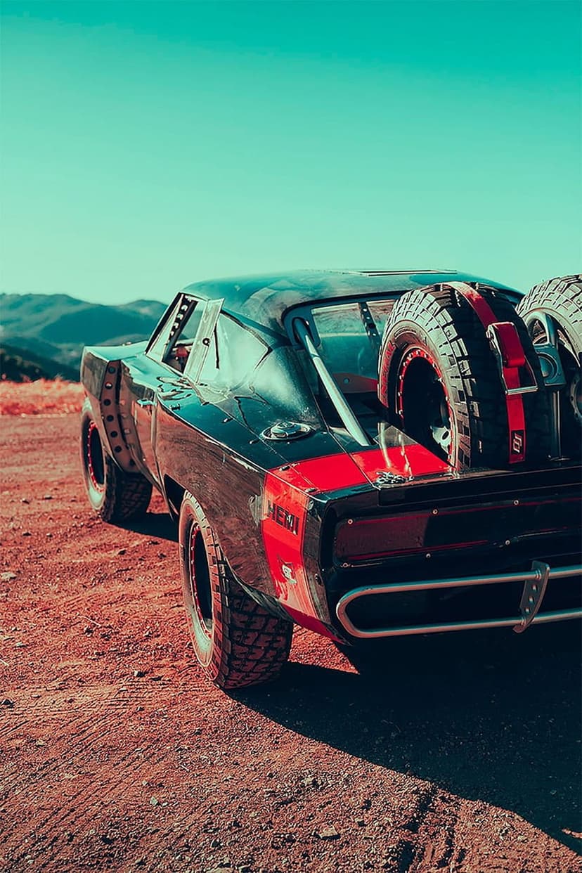 Black Dodge Charger with off-road tires and rear-mounted spares sits on a dusty desert under a blue sky.