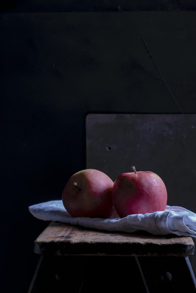 Red apples rest on a white cloth and rustic wooden stool against a dark, textured, moody background.