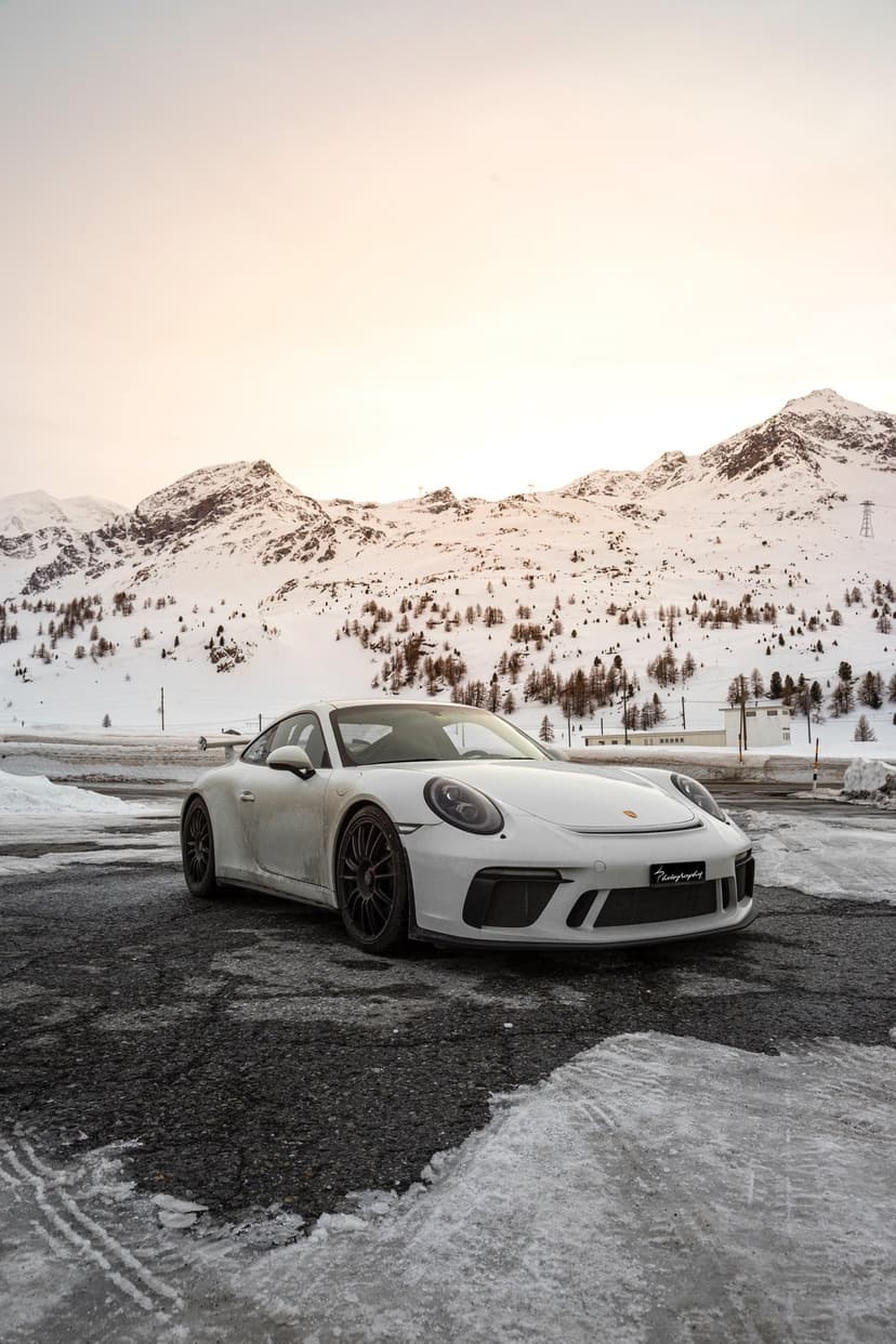 White Porsche 911 sports car parked on a snowy mountain road with hazy peaks and a winter sky in the background.