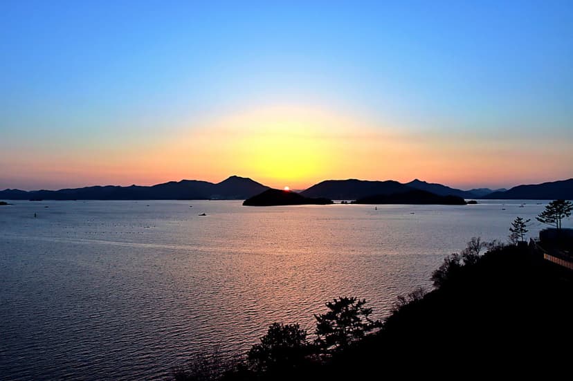 Silhouetted tropical islands rest on a calm sea beneath a vibrant orange sunset framed by dark shoreline leaves.