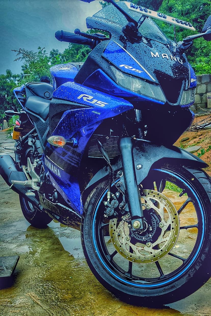 Yamaha R15 motorcycle in blue parked on wet asphalt with rain reflections and greenery under a cloudy sky.