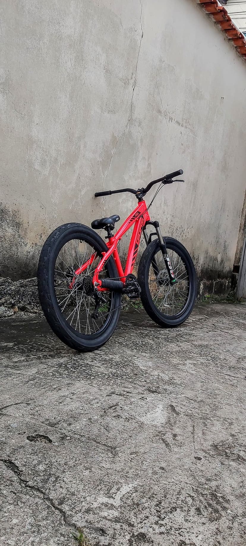 Red mountain bike leans against a gray textured wall with black tires and handlebars on a paved urban ground.