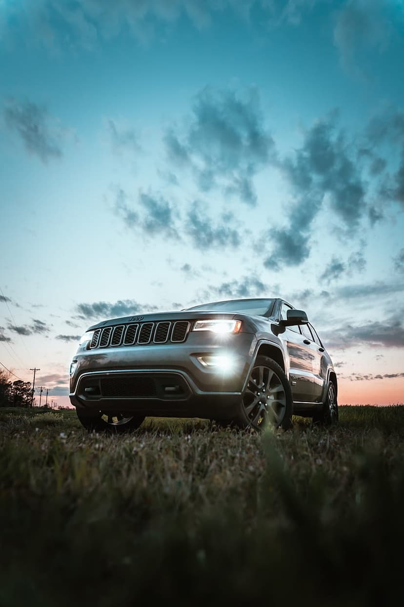 Jeep Grand Cherokee with lit headlights parked in a grassy meadow under a dramatic orange and blue sunset sky.