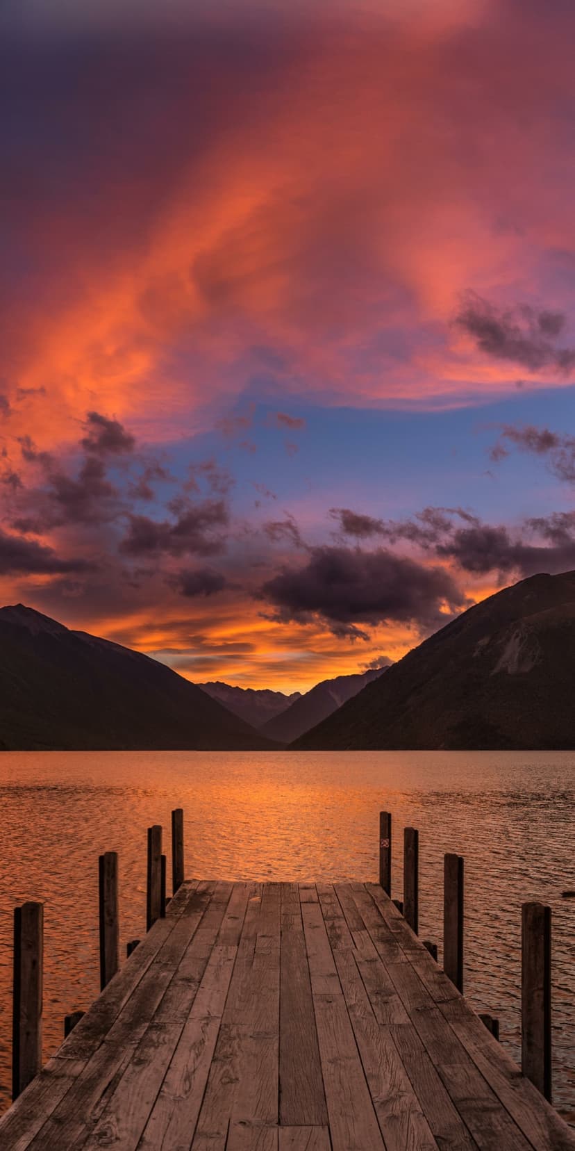 Weathered wooden dock extends into a still lake reflecting a vibrant orange sunset sky and mountain silhouettes.