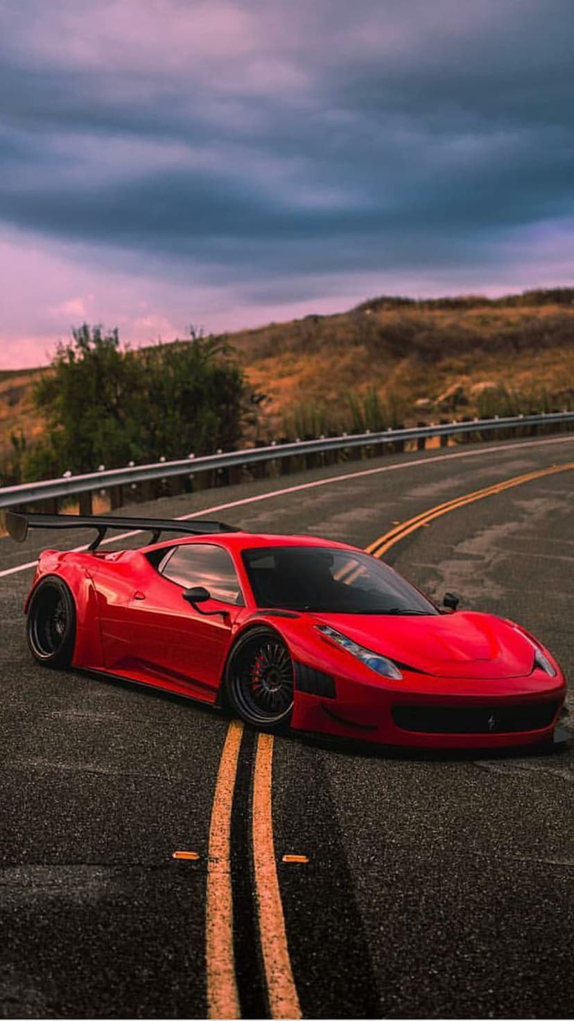 Red Ferrari with a large rear wing drives on a winding road with yellow lines under a purple and orange dusk sky.
