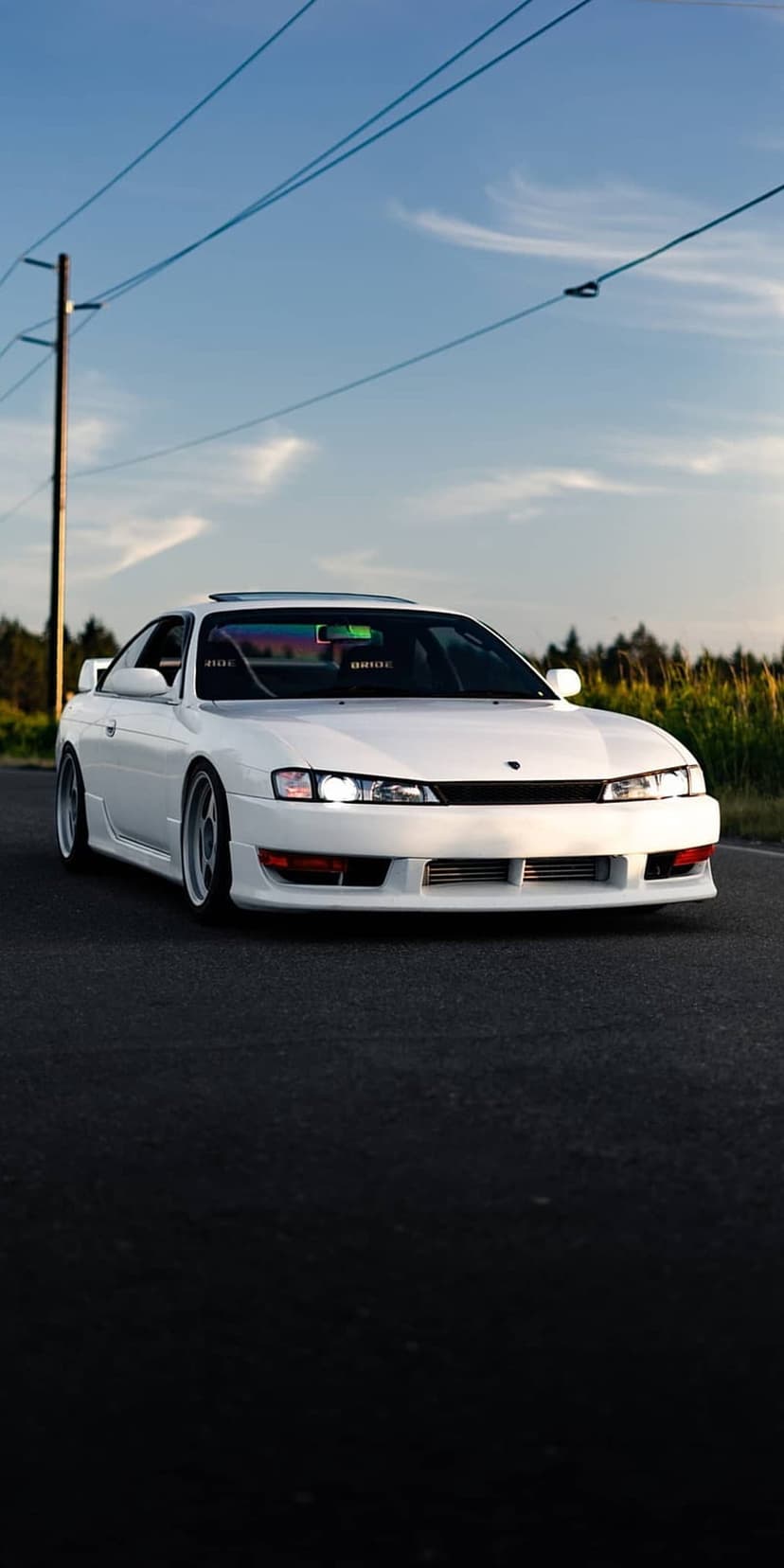 White Nissan Silvia S14 coupe on asphalt beneath power lines and a bright sky with scattered white clouds.