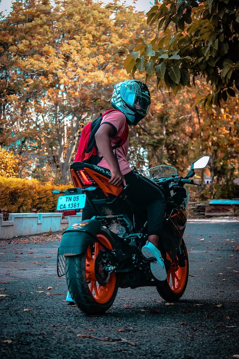 Motorcyclist with a red backpack sits on an orange bike surrounded by trees with orange and yellow autumn leaves.