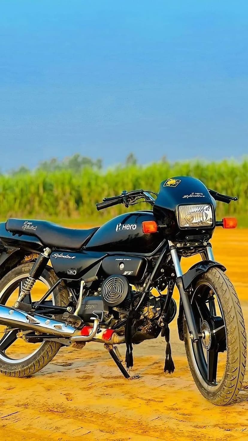 Black Hero Splendor bike parked on a dirt trail with chrome accents and green foliage under a clear sky.