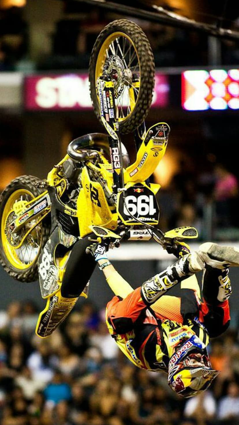 Motocross rider in yellow and orange gear performing a backflip on a dirt bike above a blurred arena crowd.