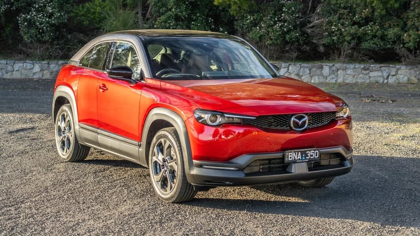Red Mazda MX-30 crossover SUV parked on a gravel path with a forest background in warm golden hour light.