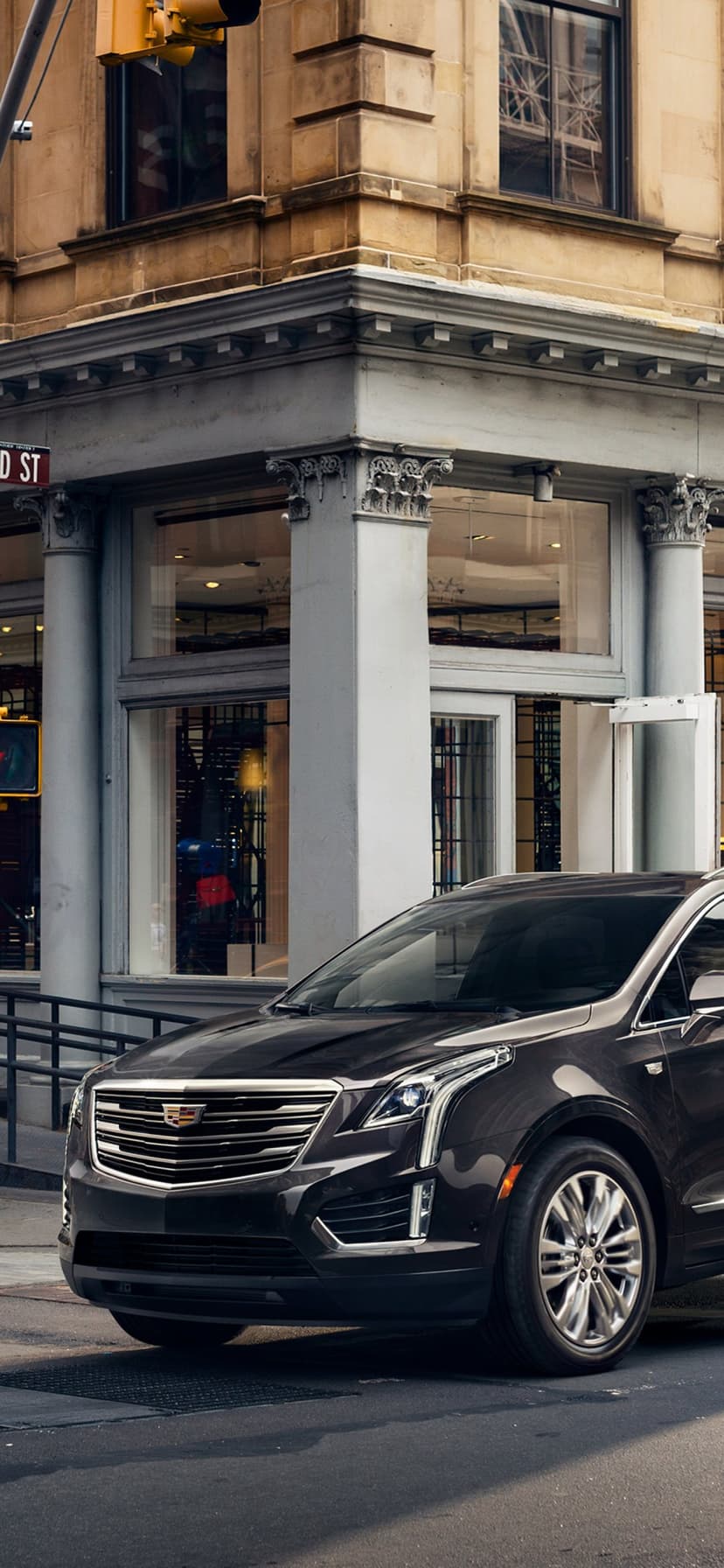 A dark brown Cadillac SUV parked on a city street corner in front of a historic classical stone building.