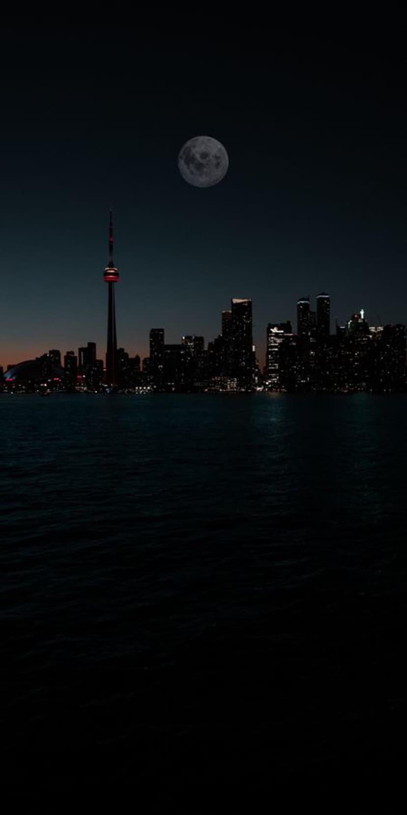 Skyscrapers and a glowing tower stand under a large full moon reflected in the calm harbor water at night.