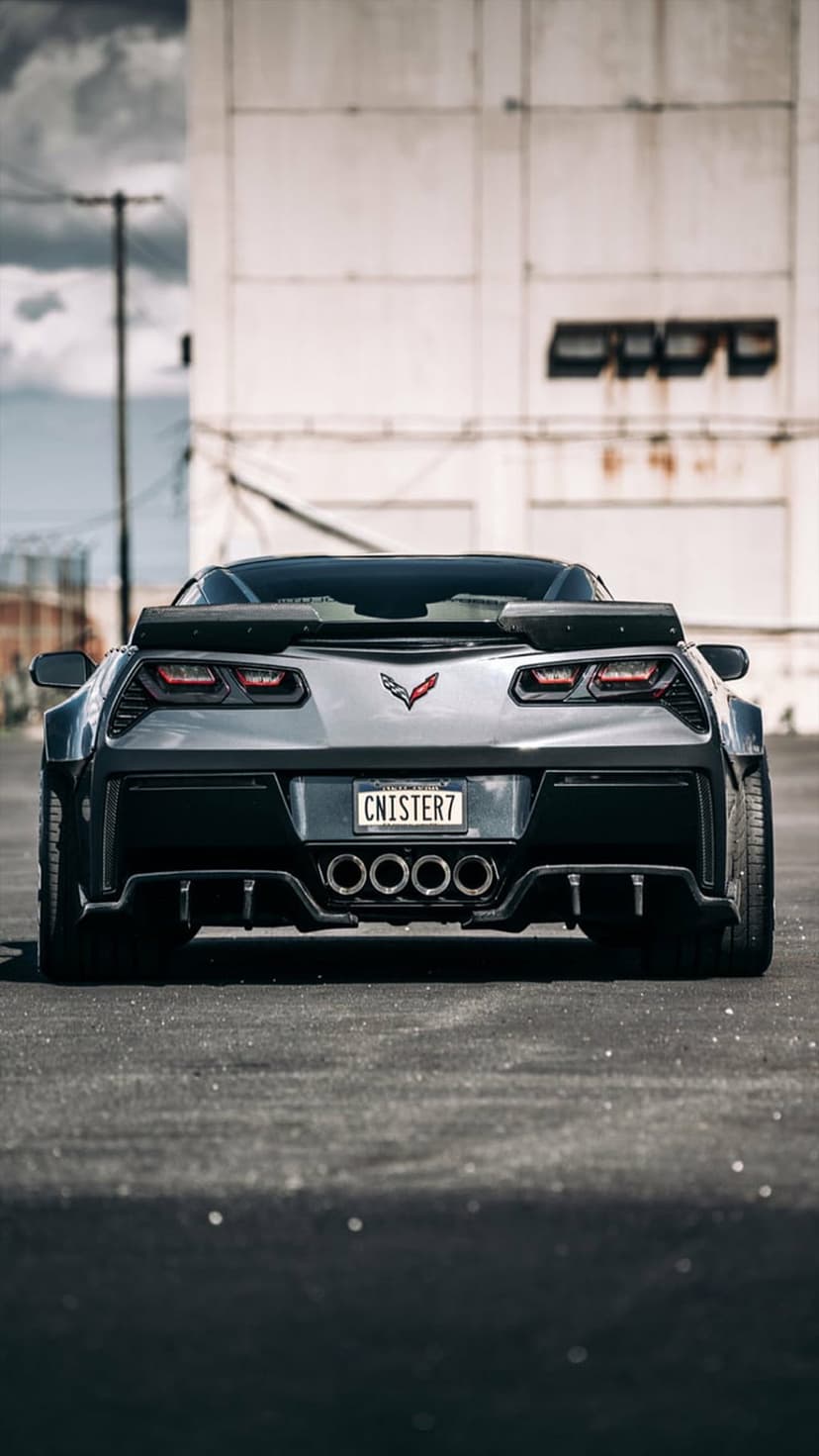 Corvette C7 Z06 rear with a large spoiler and quad exhaust parked on asphalt by a weathered industrial building.