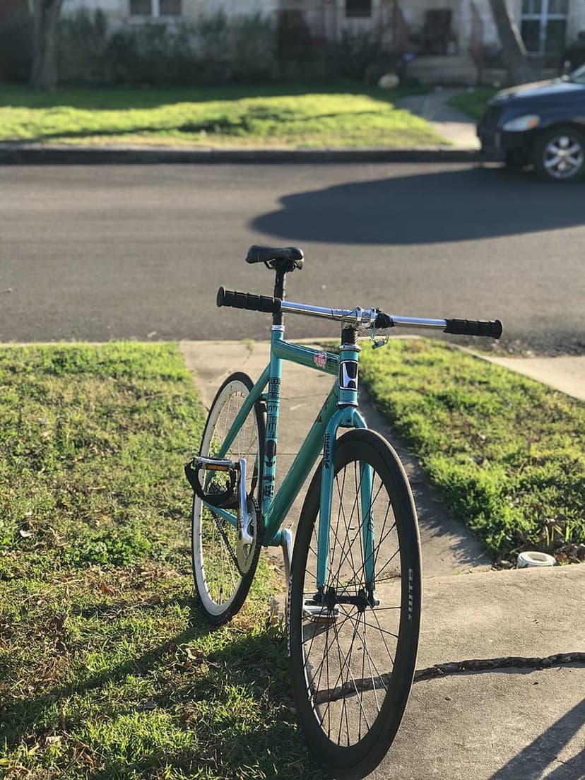 Teal fixie bicycle with drop handlebars parked on a sunlit sidewalk next to green grass and a city street.