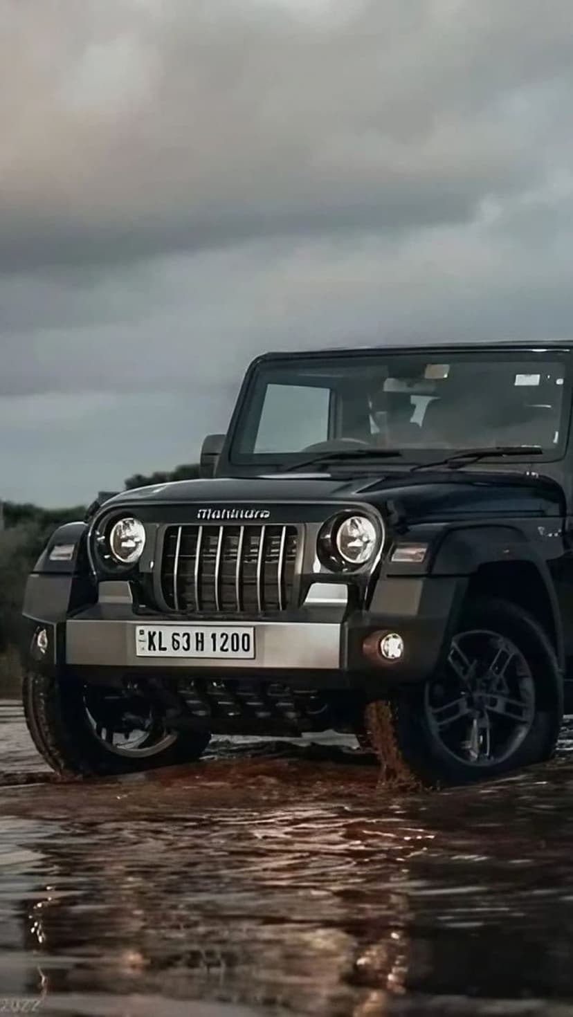 Black Mahindra Thar SUV splashing through shallow muddy water under a dramatic and moody overcast sky.