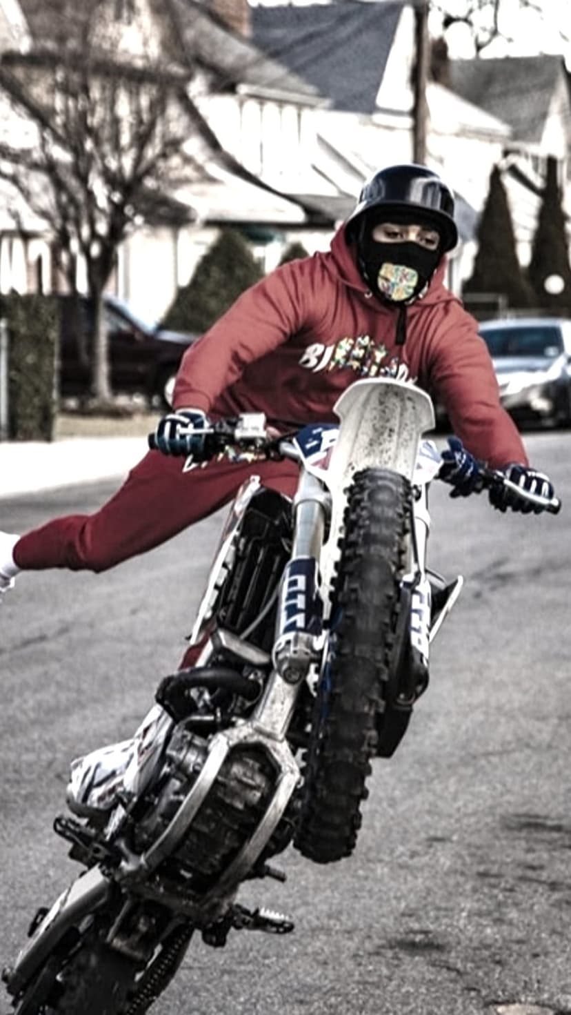 Motorcyclist in a red hoodie performs a high wheelie on a blue and white dirt bike in a residential neighborhood.
