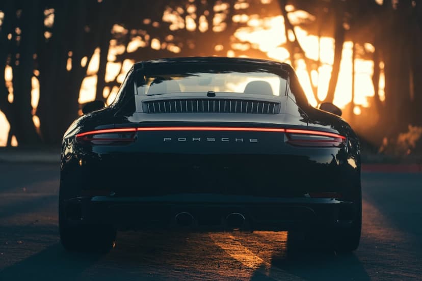 Rear view of a black Porsche 911 at sunset with glowing red light bar and sunlit trees in the background.