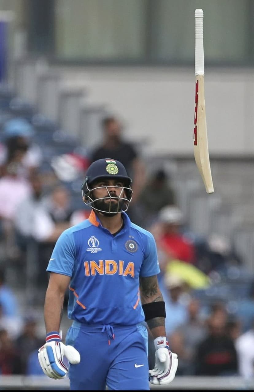 Virat Kohli in a blue jersey looks up as his cricket bat flies mid-air during a match with a blurred crowd behind.