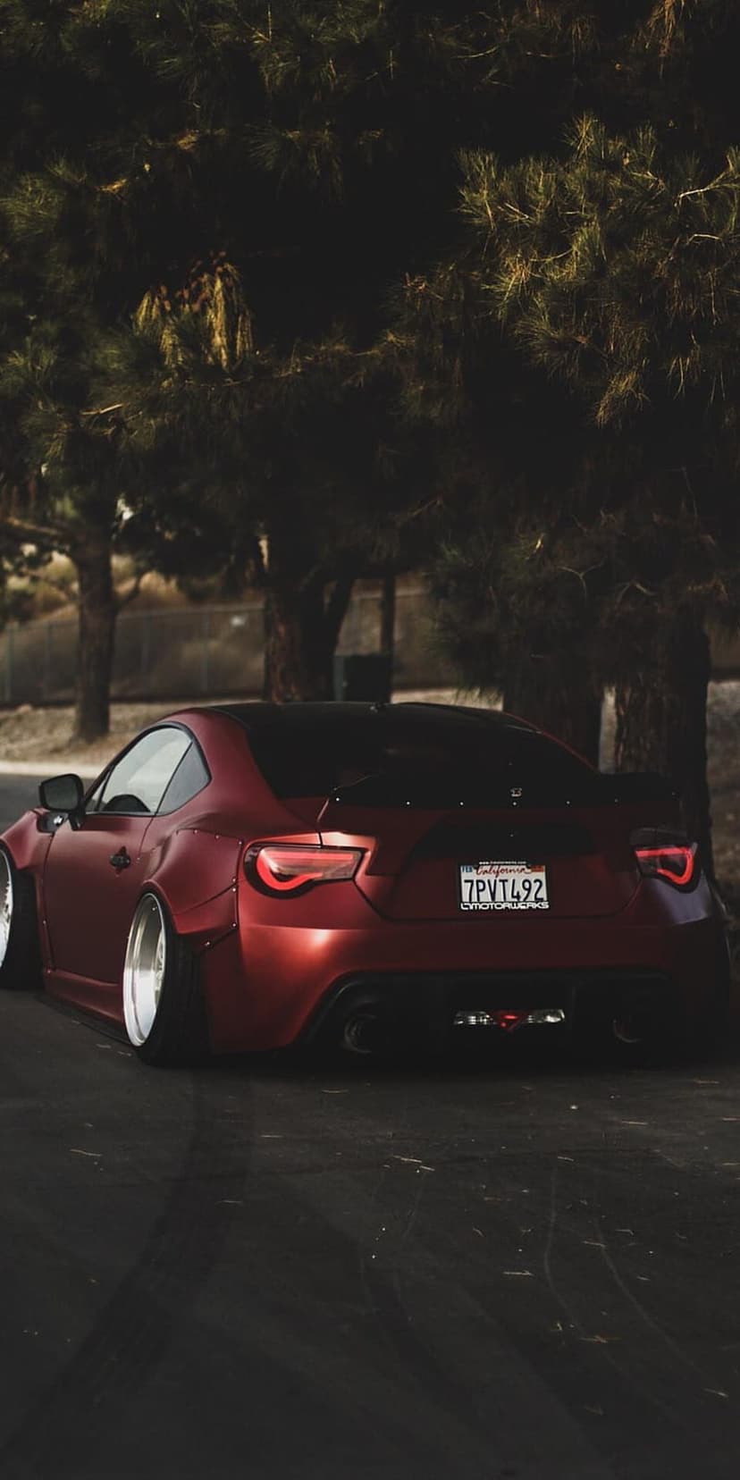 Matte red widebody sports car with custom taillights parked against a dark tree line on a vertical mobile screen.