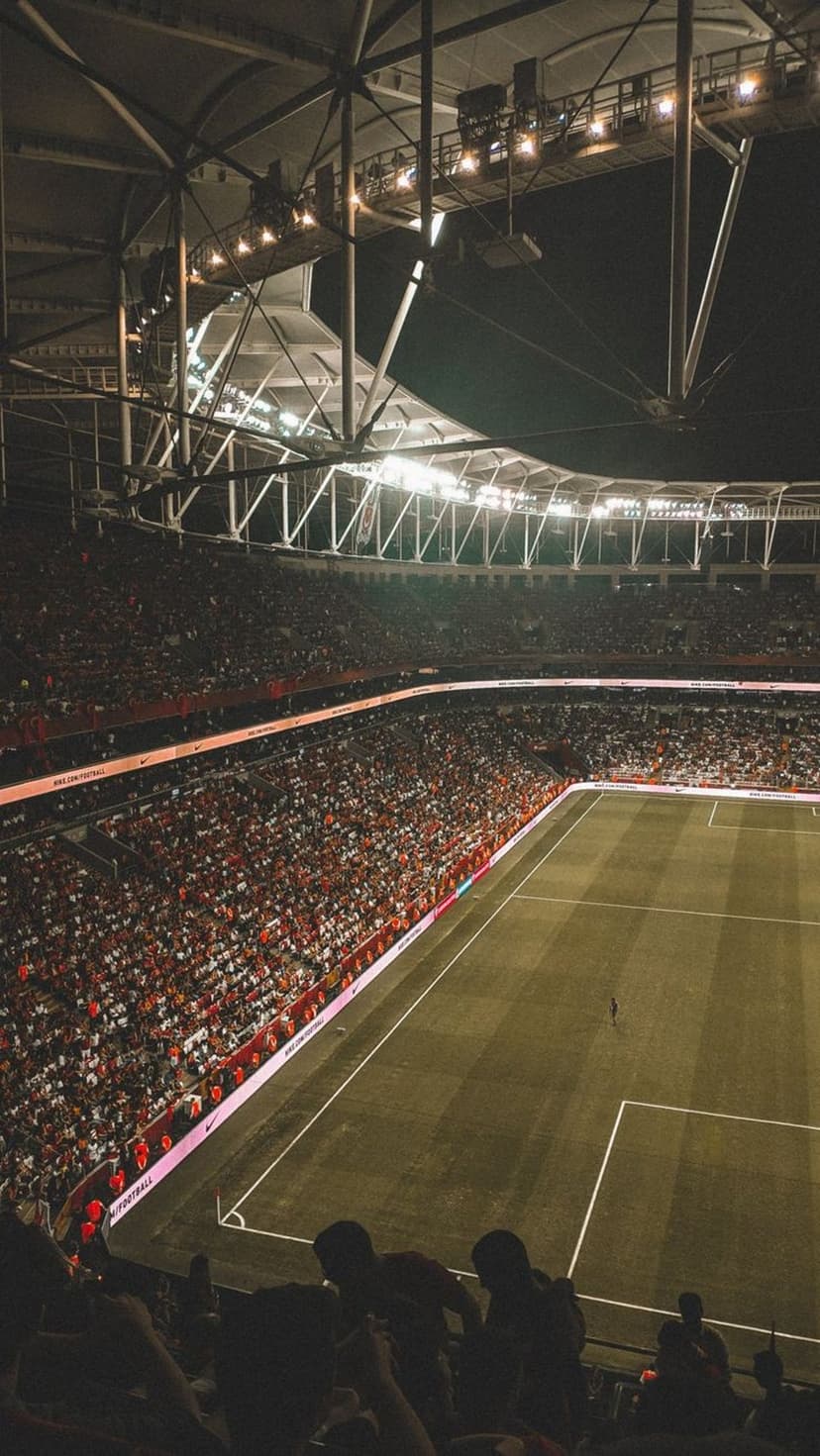 Soccer stadium glowing under night lights with red and white crowds filling the stands from an aerial view.
