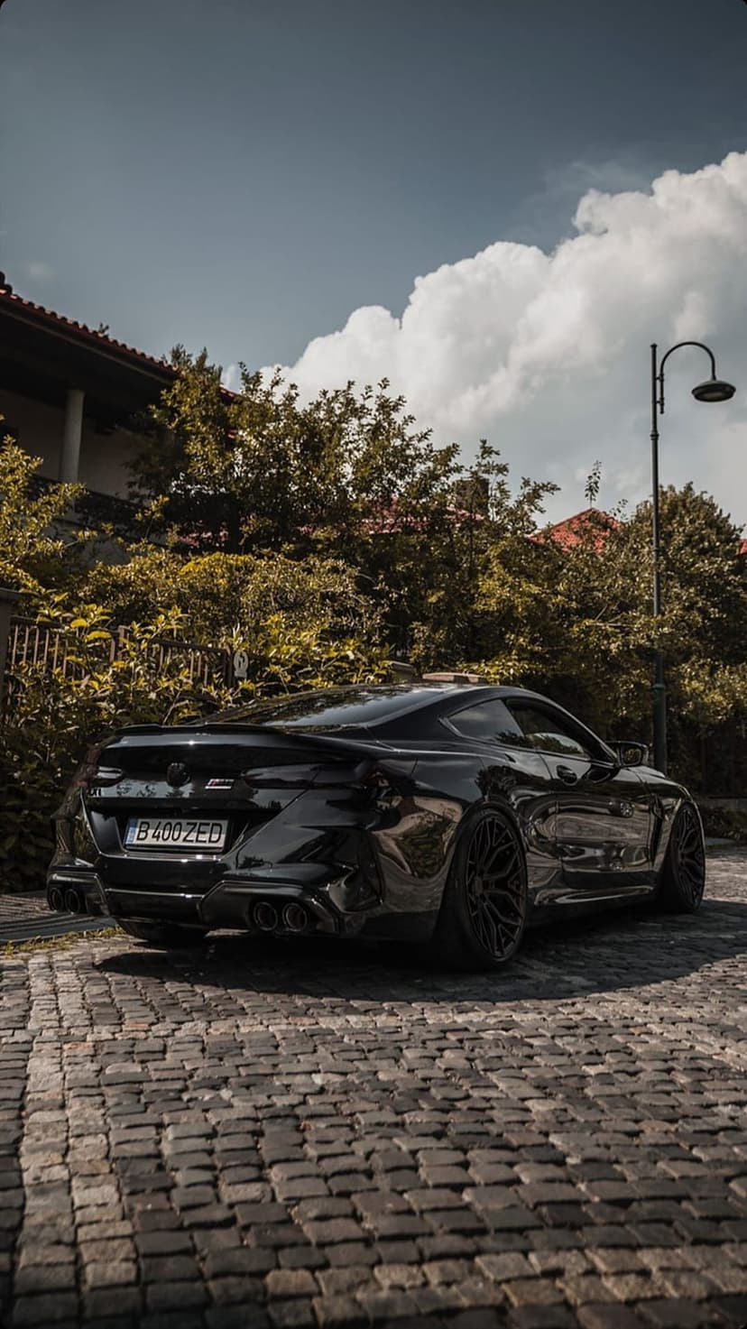 Black BMW M8 Coupe showing aggressive rear exhausts and wheels on a cobblestone road near a leafy green tree.