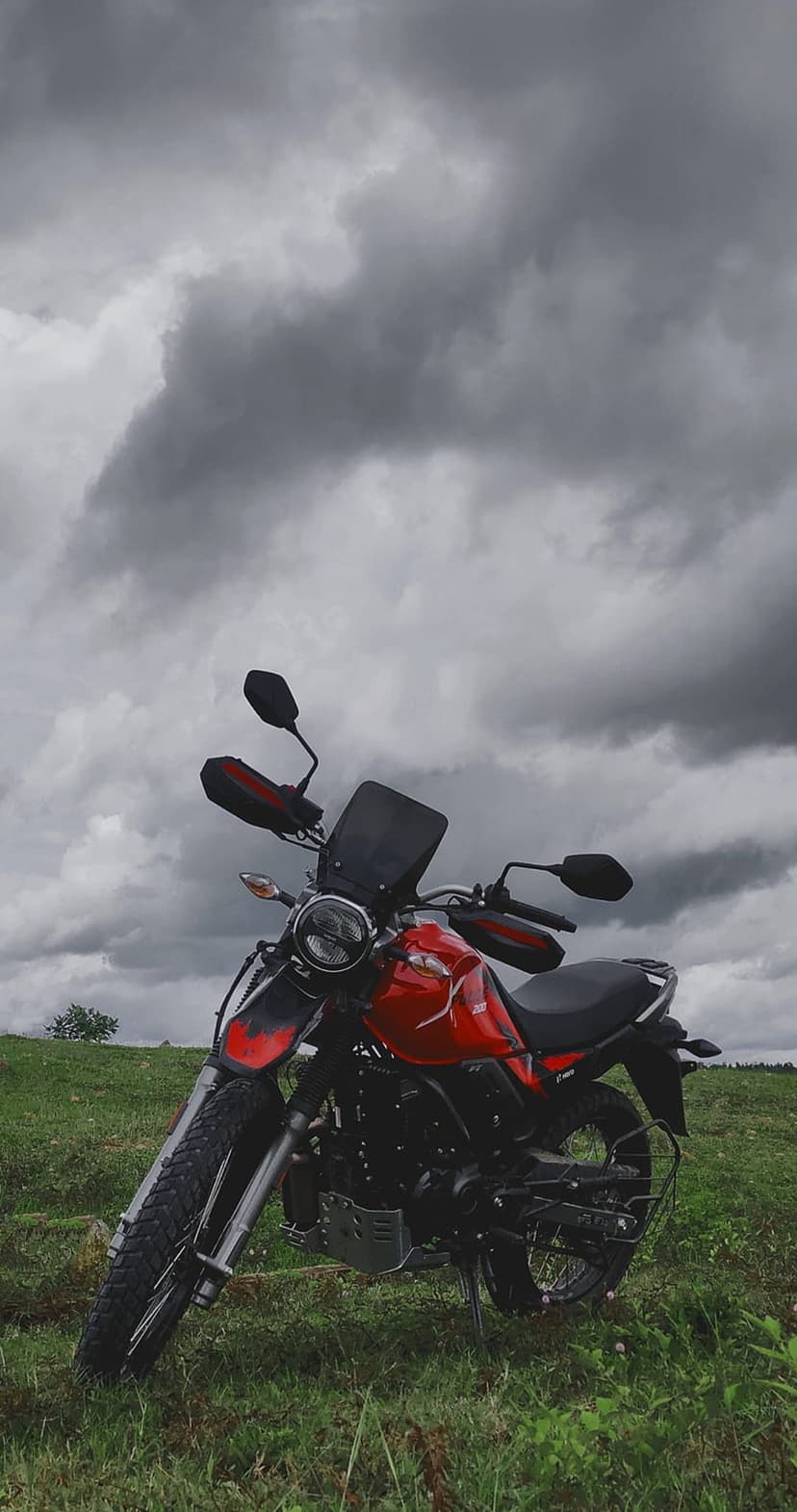 Red adventure motorcycle parked on a lush green hill under dark, dramatic storm clouds in a rural landscape.