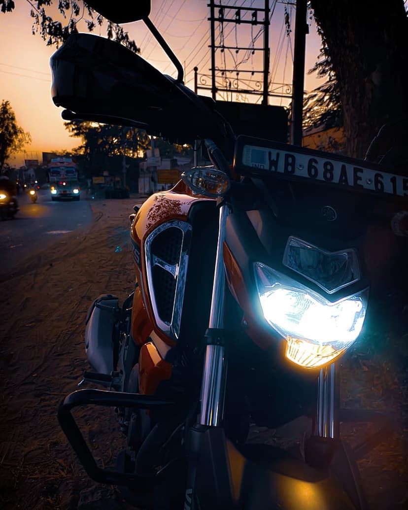 Glowing motorcycle headlights on an orange bike shine onto a road with a sunset and silhouettes in the distance.
