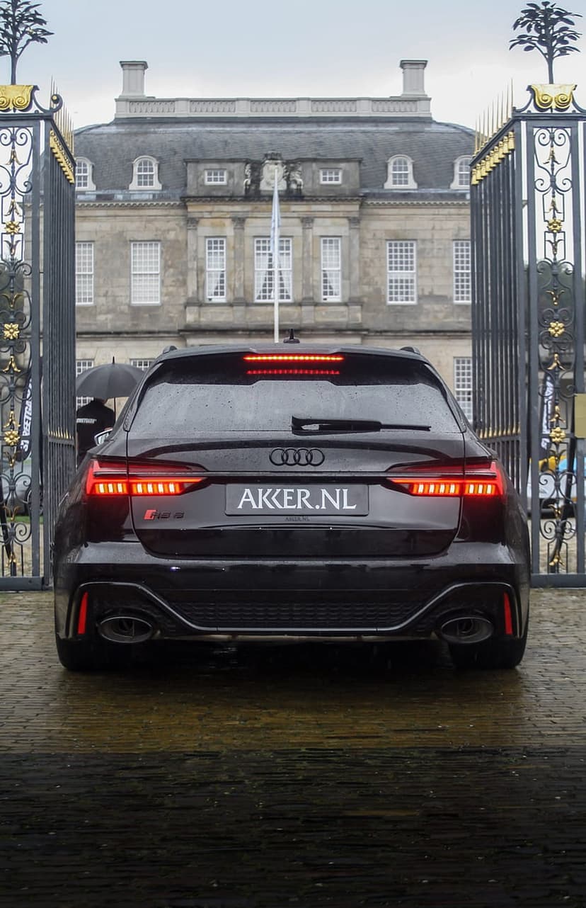 Audi RS6 Avant rear view on wet cobblestones before a gold-accented gate and historic stone manor house.