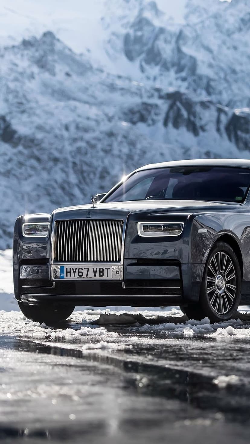 Charcoal Rolls-Royce Phantom parked on a snowy road with sunlit headlights and jagged mountain peaks behind it.