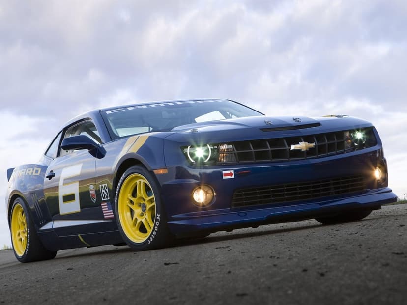 Blue Chevrolet Camaro race car with yellow wheels and illuminated headlights on an asphalt track under cloudy skies.