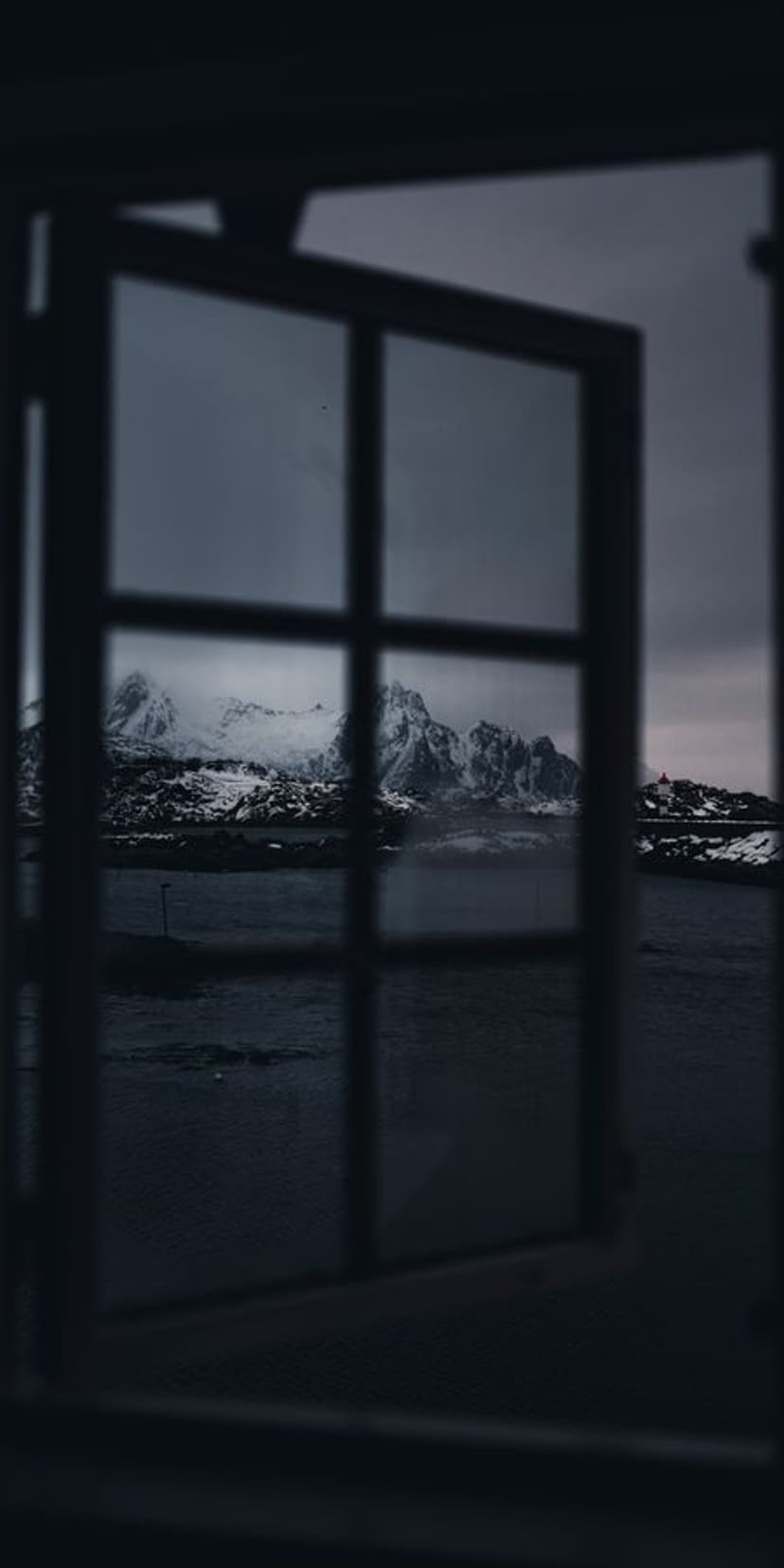Snow-capped mountains and a red cabin seen through a dark window frame above icy, dark arctic waters.