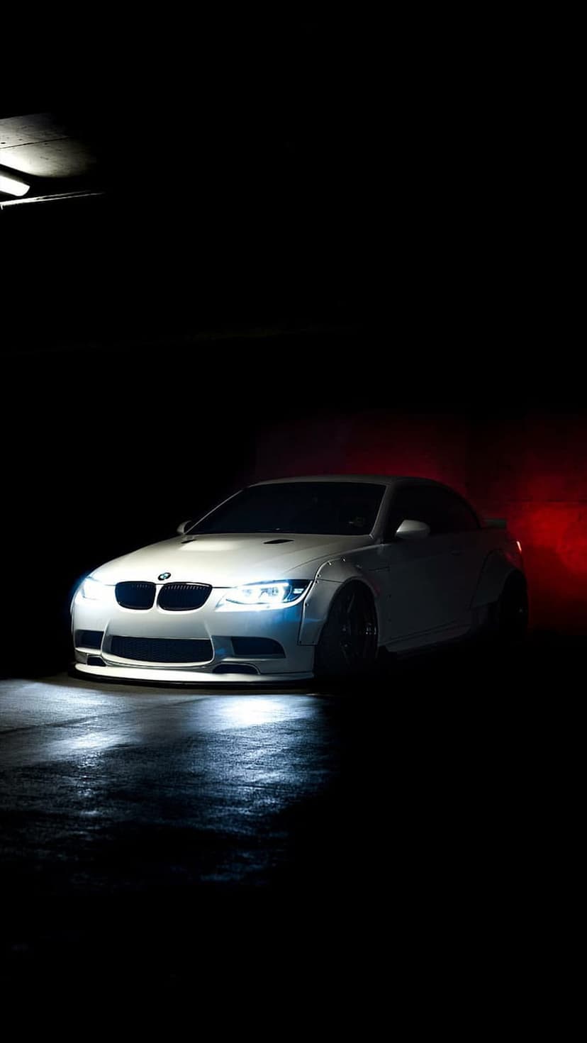 White BMW M3 coupe parked on a wet street at night with glowing red background lights and bright LED headlights.
