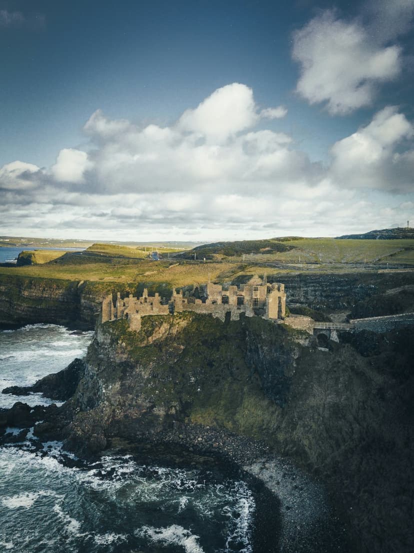 Dunluce Castle ruins stand on a rocky cliff edge above churning ocean waves and a green coastline in Ireland.