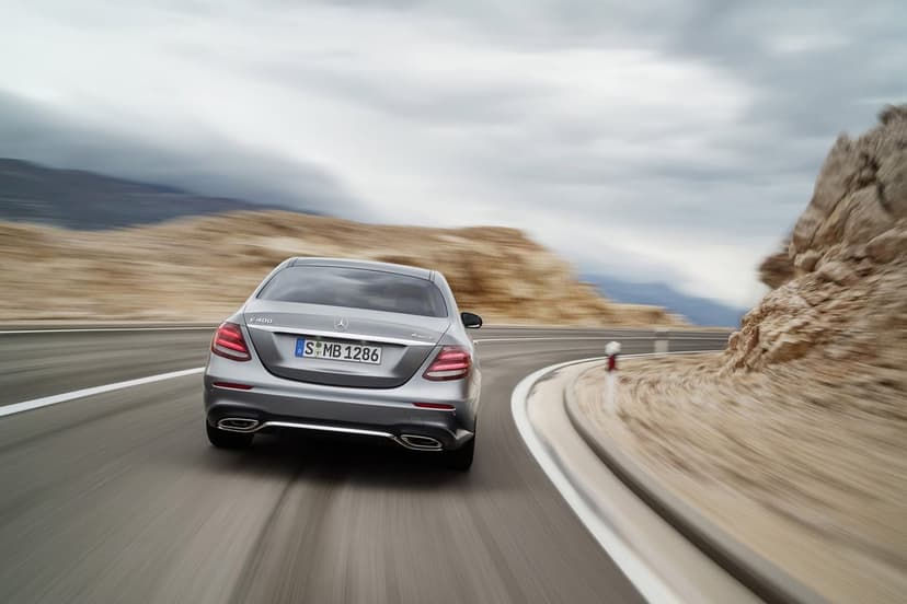 Silver Mercedes-Benz sedan driving around a coastal bend with motion blur and rocky landscape background.