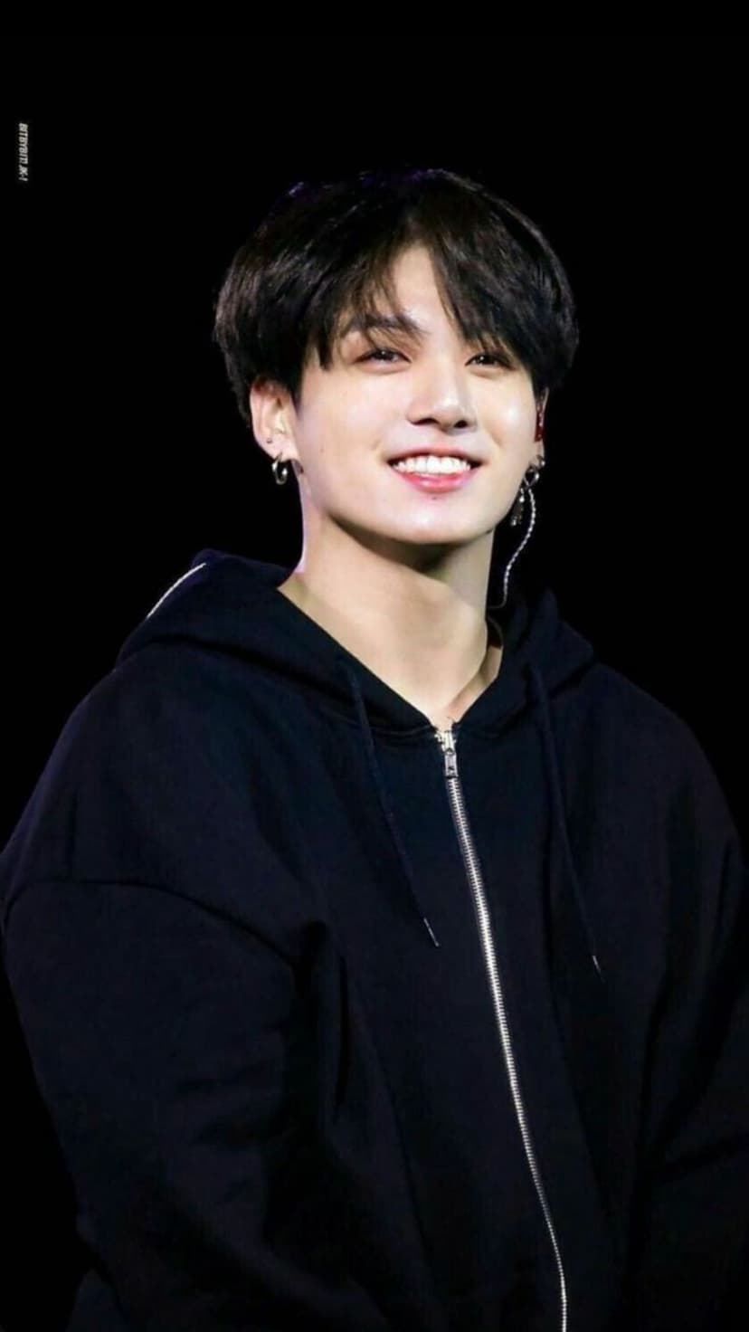 Jungkook from BTS smiles warmly while wearing a black zip-up hoodie against a high-contrast dark background.