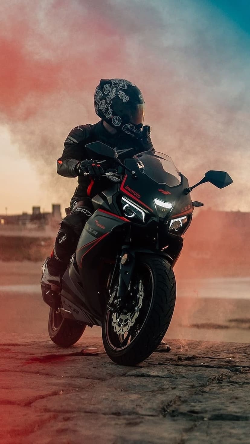 Motorcyclist in full gear on a black sportbike sits amidst swirling red and blue smoke on a coastal road at dusk.