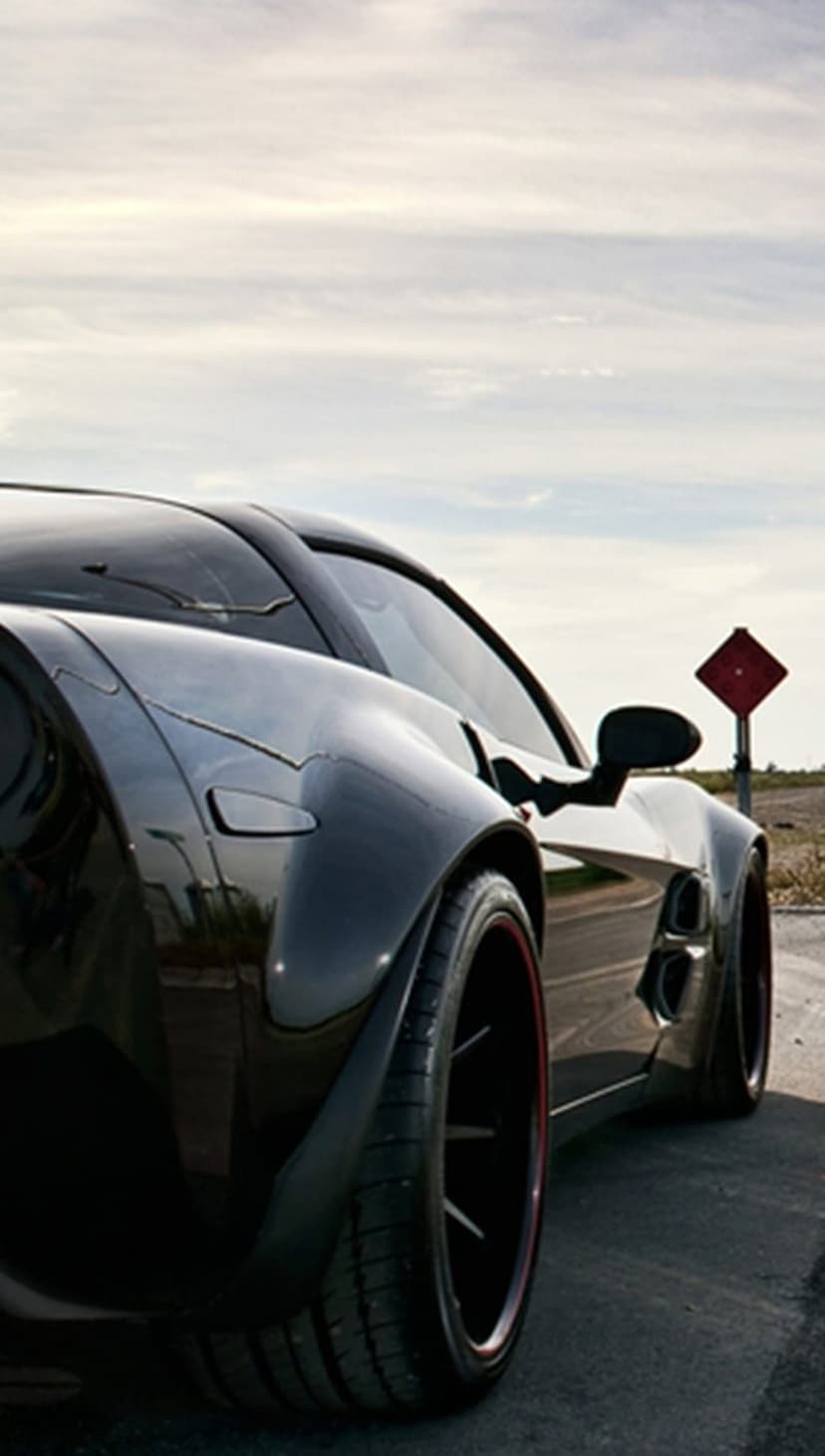 Low-angle black sports car featuring custom red rims parked on a gray asphalt road under a bright clear sky.