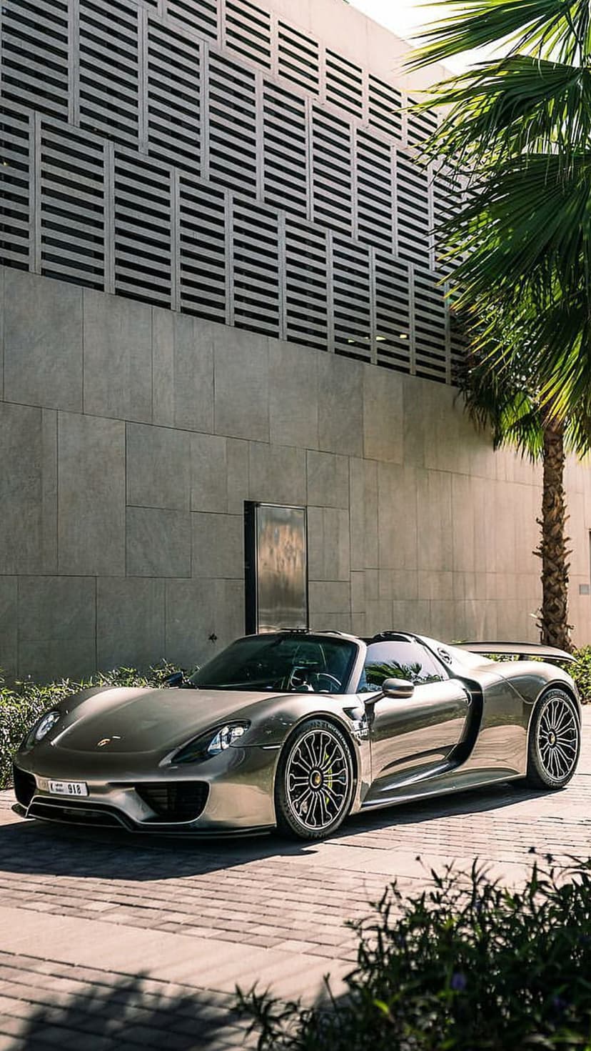 Silver Porsche 918 Spyder parked on brick pavers beside a tall palm tree and a modern building with gray louvers.