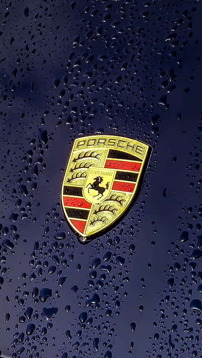 Porsche crest emblem sits on a dark blue metallic car hood covered in small, clear shimmering water droplets.