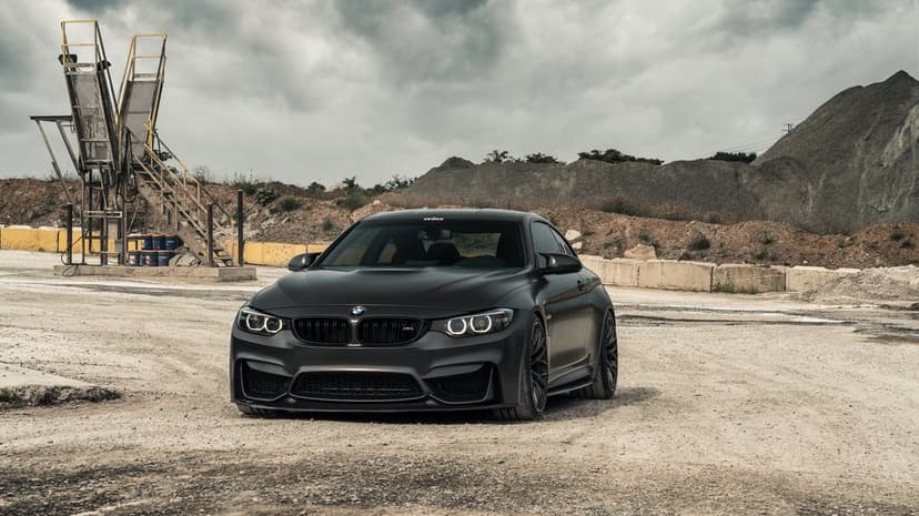 Matte black BMW M4 parked on a gravel lot with industrial mounds and a cloudy sky in the background.
