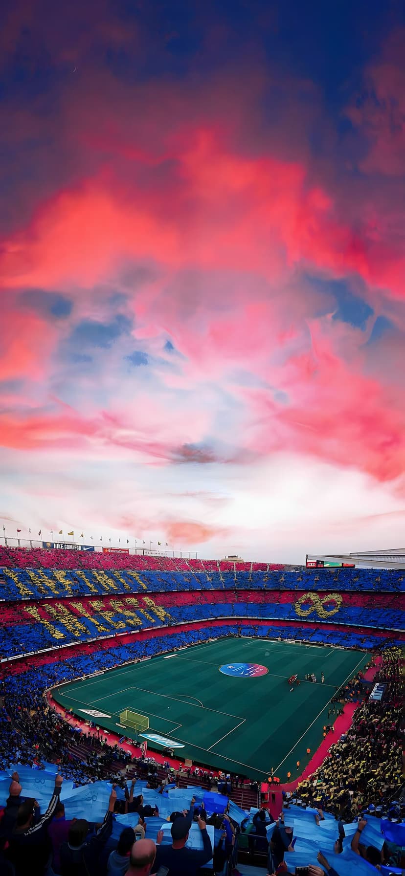 Thousands of fans create a red and blue mosaic in a large stadium under a pink and orange sunset sky at dusk.
