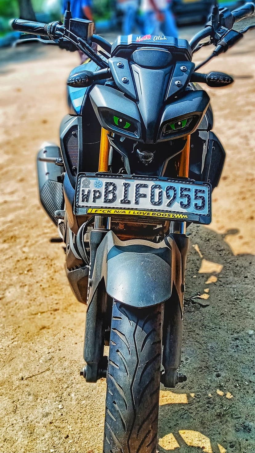 Yamaha MT-15 motorcycle with glowing green cat-eye headlights and a matte grey finish on a sandy outdoor terrain.