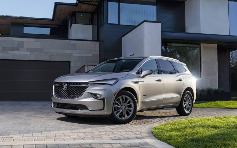 Silver Buick Enclave Avenir parked on a stone driveway before a modern house with large glass windows.