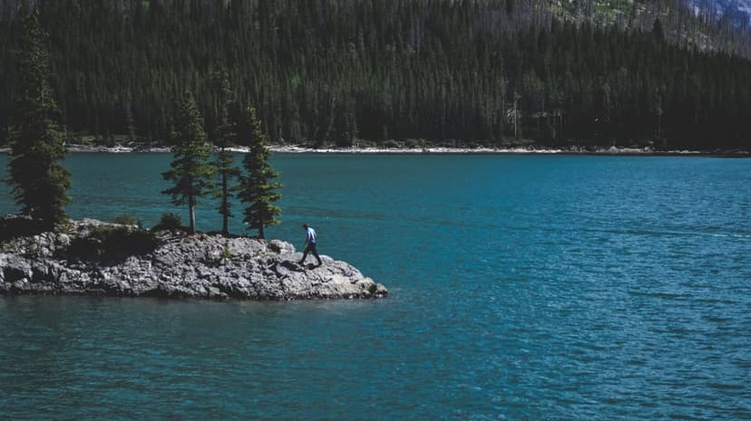 A man walks across a small rocky island with evergreen trees surrounded by vast, rippling turquoise lake water.