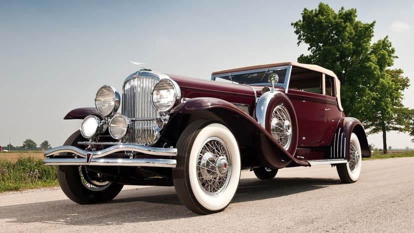 A maroon vintage luxury car with chrome accents and white-wall tires parked on a sunlit road under a blue sky.