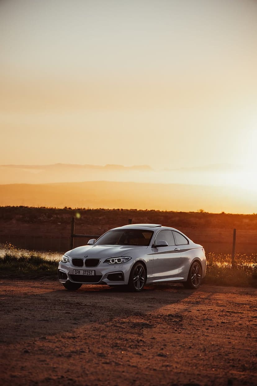 Silver BMW coupe parked on a rural dirt road surrounded by dry grass under a vibrant orange and yellow sunset sky.